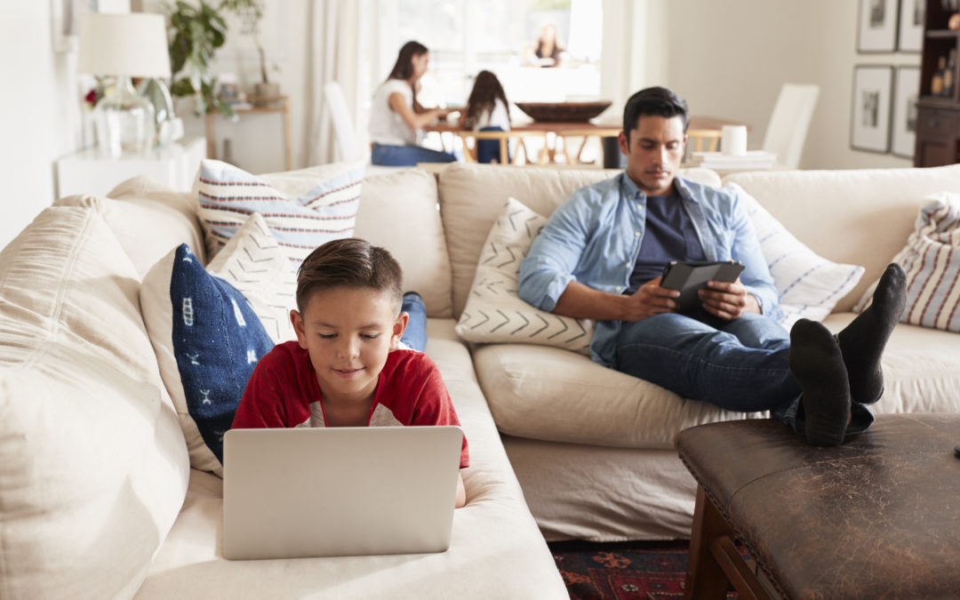 Pre-teen boy lying on sofa using laptop, dad sitting with tablet, mum and sister in the background